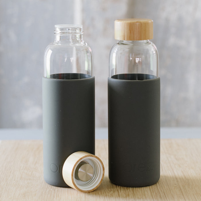 Two glass water bottles with bamboo lids are placed on a wooden surface with a grey background.
