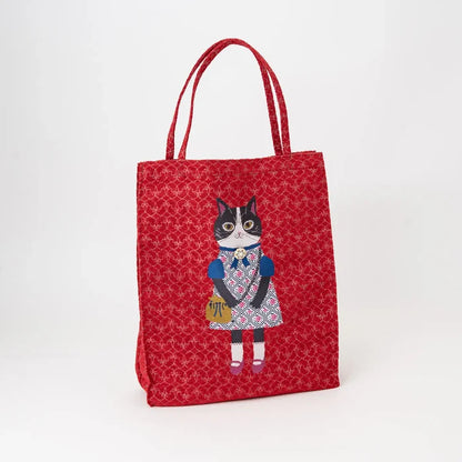 A red canvas tote bag with a repeating floral pattern and a central illustration of a black-and-white cat wearing a blue and pink dress, standing on a white background.