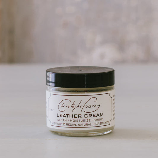 A 2oz glass jar of Leather Cream by Christophe Pourny Ltd., with a black lid, placed on a light surface.