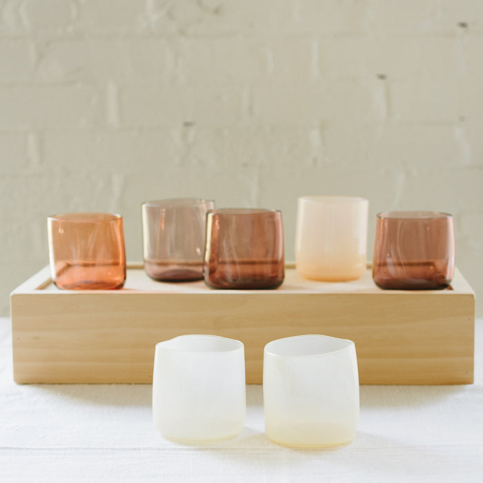 A set of various handcrafted, mouthblown glass cups displayed on a wooden tray against a brick wall background.