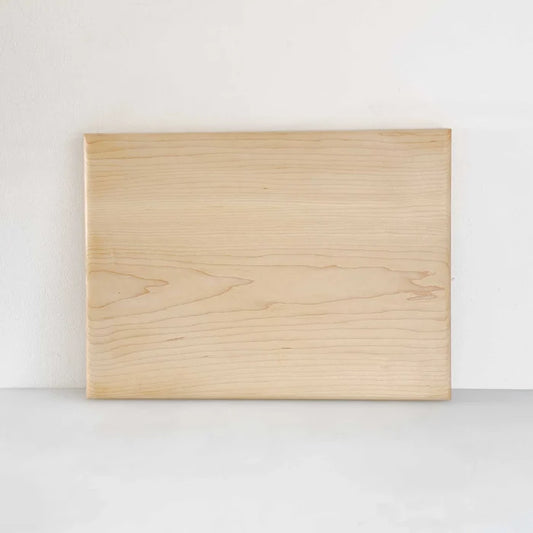 A rectangular light wood cutting board with visible natural grain patterns, standing upright against a plain white wall on a light gray surface.