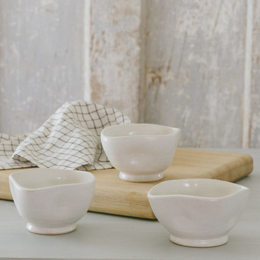 Three white stoneware bowls with white glaze on a wooden surface, with a checkered napkin in the background.